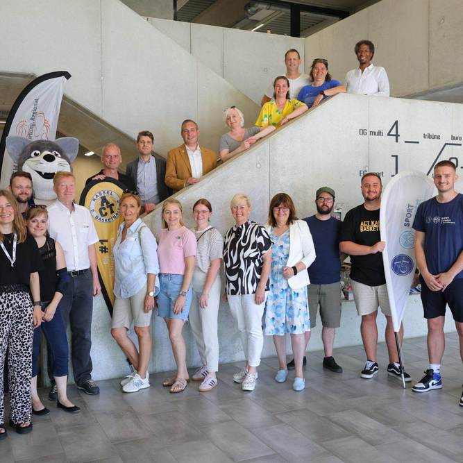 Gruppenfoto mit rund 25 Personen, zwei Maskottchen und zwei Beachflags in einem Treppenhaus, darunter Sascha Holstein, NHW-Regionalcenterleiter Kassel, Alexander Hauschild (Soziale Quartiersentwicklung NHW) und Kassels Sportdezernent Heiko Lehmkuhl